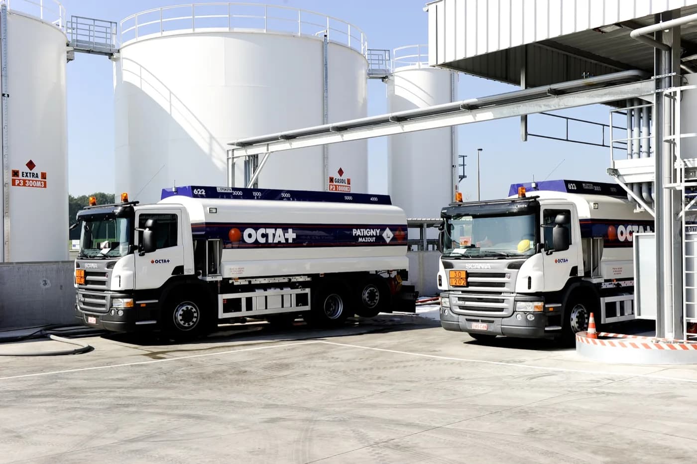 Deux camions-citernes OCTA+ stationnés devant les cuves d’un dépôt de carburant